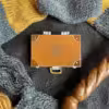 Eine Emaille-Anstecknadel mit brauner Farbe, monogrammiert mit "NS" und vor einem kuscheligen Hintergrund aus gestrickten Schals.