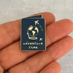 Ein anderer Blickwinkel auf die marineblaue Emaille-Anstecknadel mit dem Text "Adventure Time", die eine Person in der Hand hält.