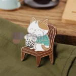 Eine Katze sitzt auf einem Stuhl neben einer Tasse Tee, im Hintergrund sind individuelle Boba-Katzenanstecker zu sehen.