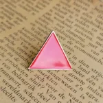 Rosa dreieckige Emaille-Anstecknadel mit der Aufschrift "Triangle Rouge", die auf einer aufgeschlagenen Buchseite ruht.