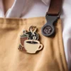 Ein Barista in einer Schürze hält eine Kaffeetasse, auf der eine schwarz-weiße Anstecknadel mit dem Barista-Abzeichen prangt.
